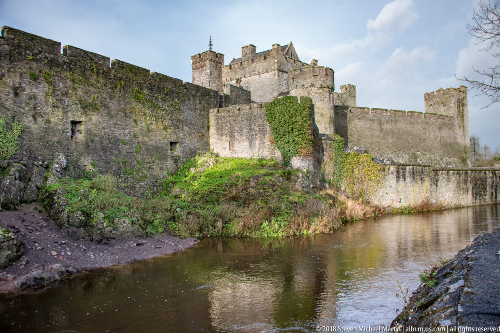 Layered stone walls and towers of Cahir Castle rising above the River Suir, with ivy on the lower masonry.
