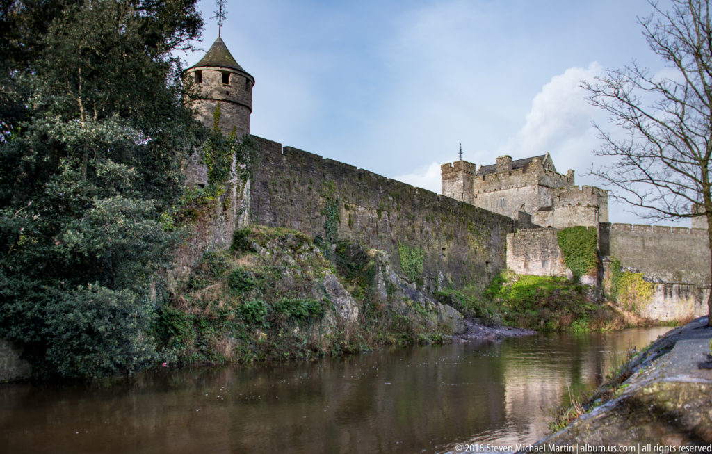 Long stone curtain wall of Cahir Castle above the river, with a roofed turret and taller inner towers.