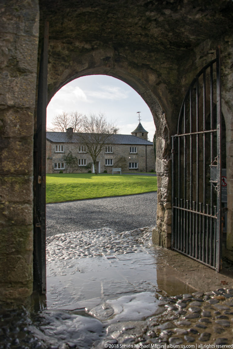 Stone archway and open iron gate framing a wet courtyard and long low building inside Cahir Castle.