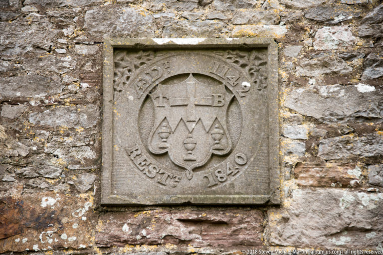 Square carved stone plaque with a cross, initials, geometric symbols, and dates set into rough masonry.