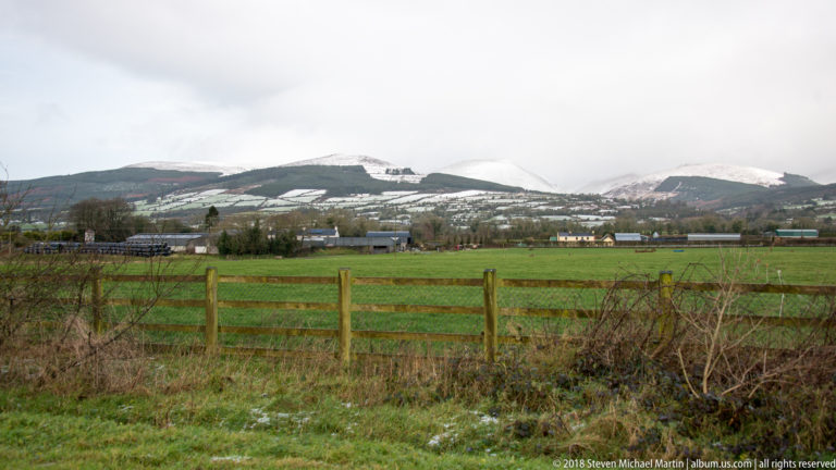 Green pasture and wooden fencing in front of farm buildings and snow-dusted hills near Cahir.