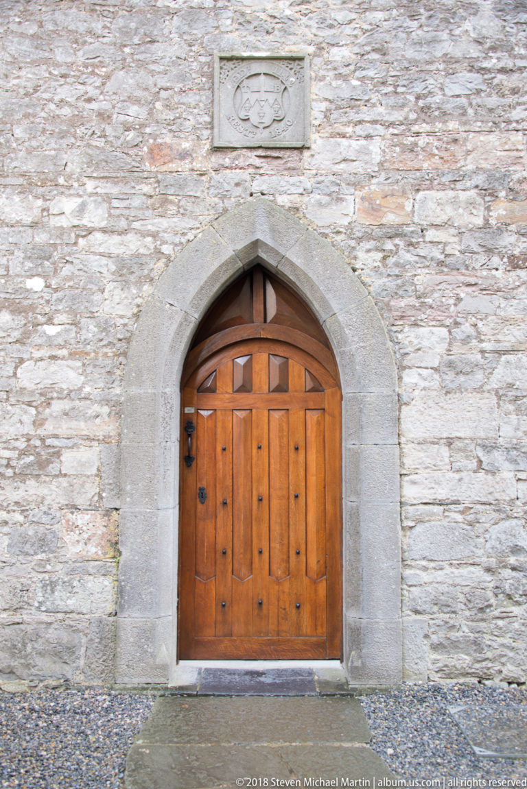 Paneled wooden door with iron hardware in a pointed dressed-stone arch set into a pale rubble wall.