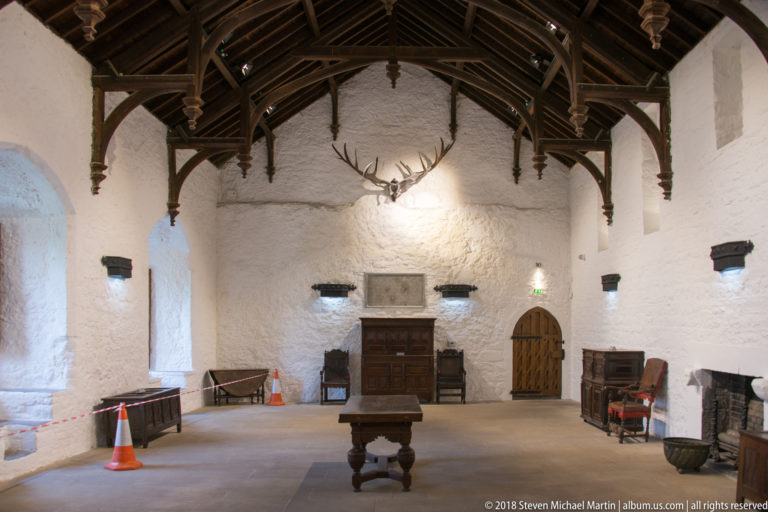 Whitewashed castle hall with an elaborate dark timber roof, historic furniture, central table, and mounted antlers.