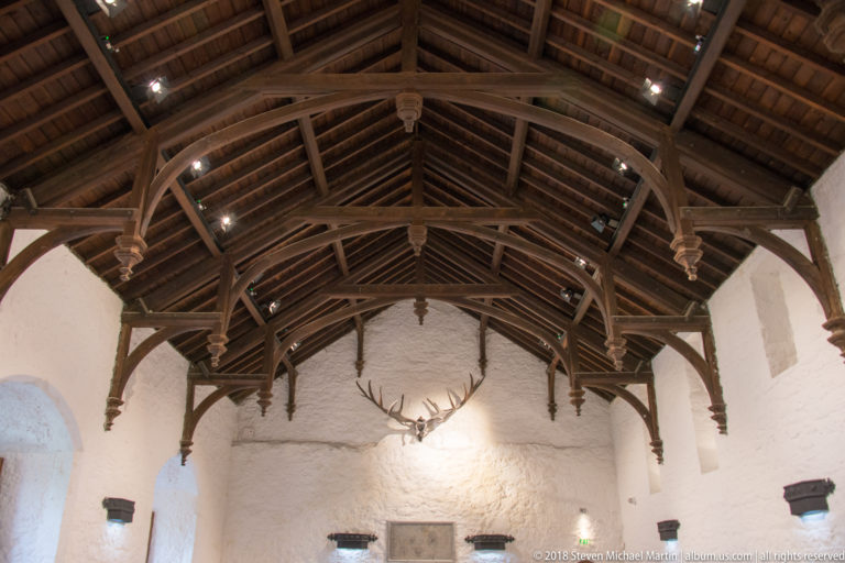 Upward view of a dark open timber roof with arched braces, carved pendants, and rafters above white walls.