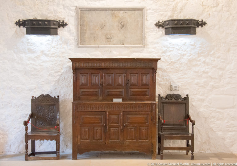 Large carved wooden cupboard between two high-backed chairs against a whitewashed wall with rope barriers.