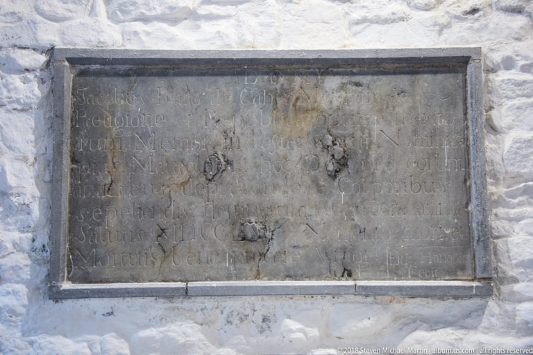Weathered rectangular stone inscription with faint carved lettering set into a whitewashed wall.