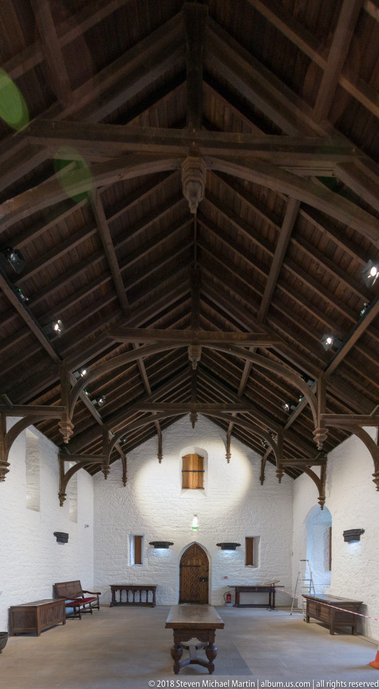 Long whitewashed hall beneath a dark timber roof, with benches, central table, small windows, and arched doorway.