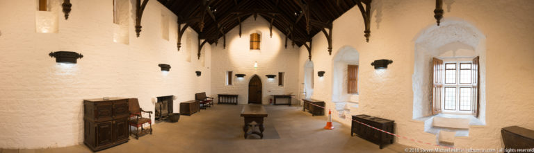 Oblique view down a whitewashed castle hall with deep windows, timber roof brackets, furniture, and a central table.