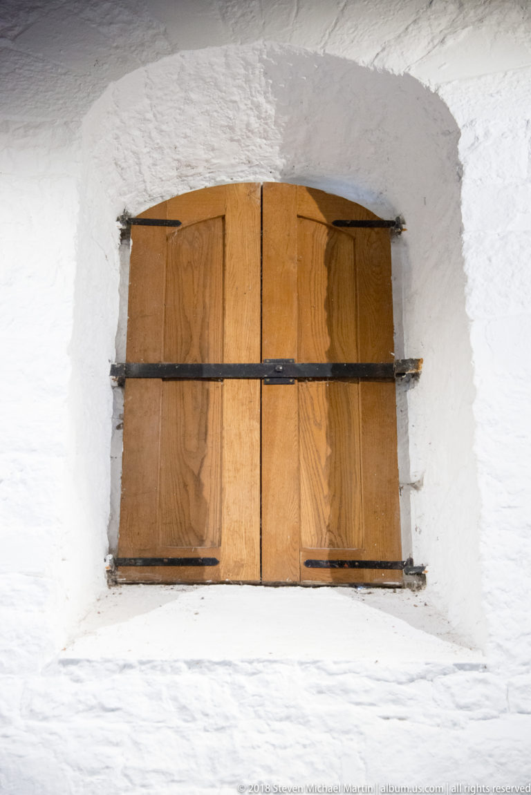 Closed wooden shutters with black iron strap hinges set deep within a thick whitewashed stone opening.
