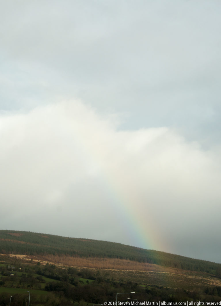 Faint rainbow descending from a gray sky above dark tree cover and low rural fields.
