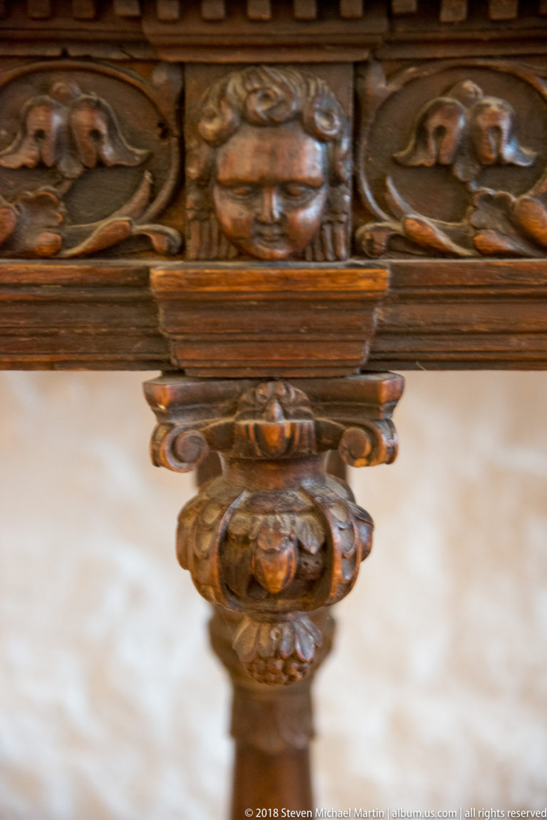 Close carved wooden furniture detail with a human face, scrolling ornament, and rounded decorative forms.