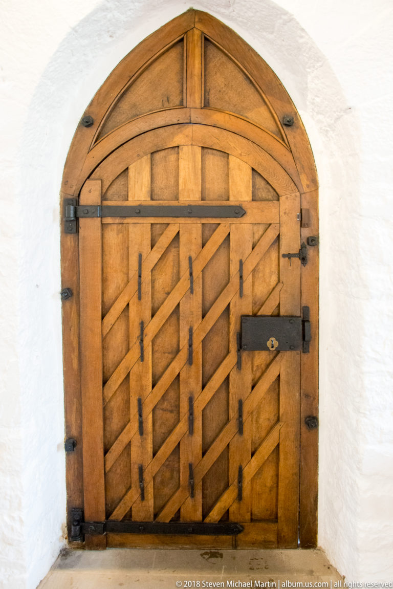 Arched wooden door with diagonal bracing, iron strap hinges, and a heavy horizontal locking bar.