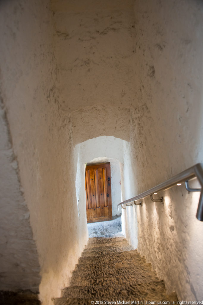 Steep stone stairs in a narrow whitewashed castle passage with handrail, small lights, and a shuttered opening.