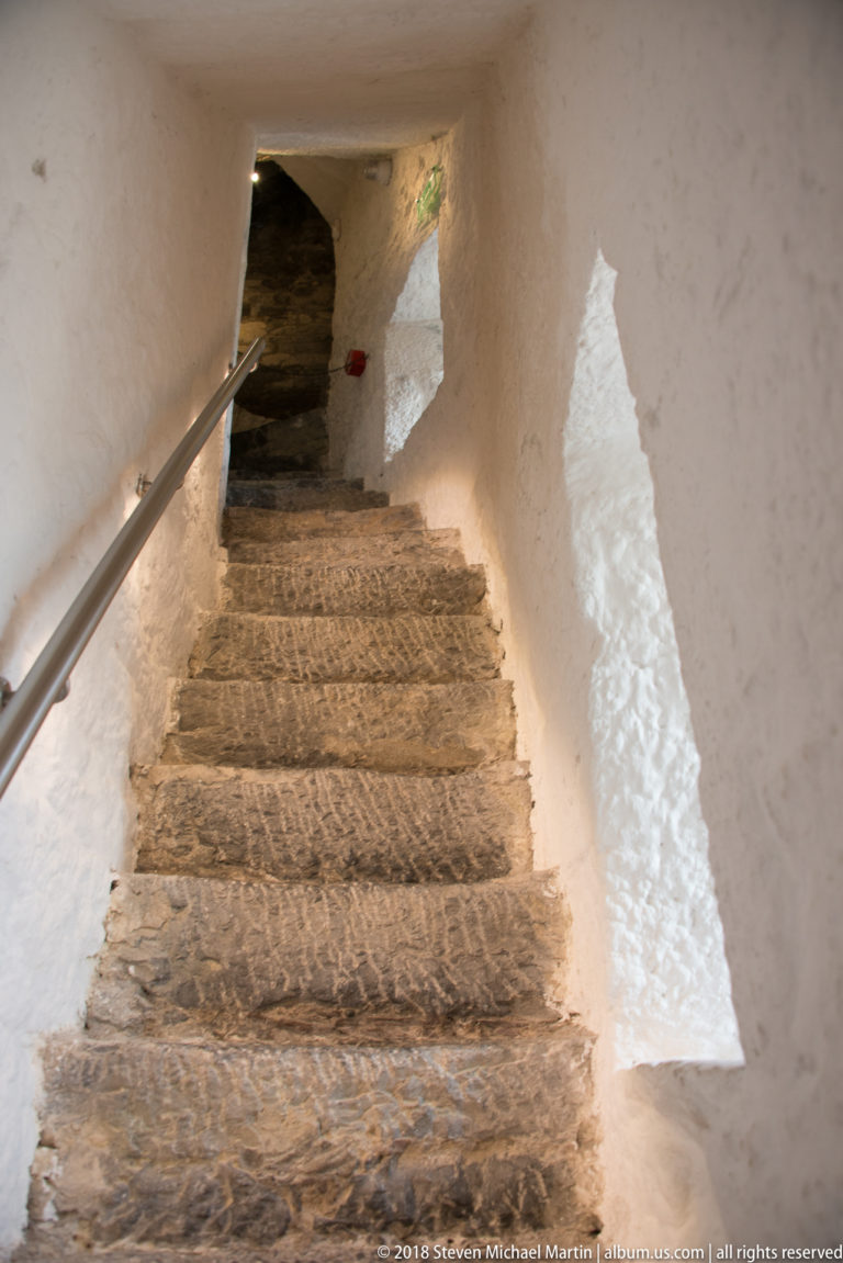 Rough stone staircase rising through a narrow whitewashed passage beside a slim vertical wall opening.
