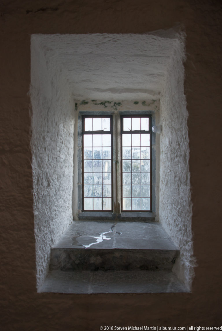 Small multi-pane window recessed deeply in a thick whitewashed castle wall above a broad stone sill.