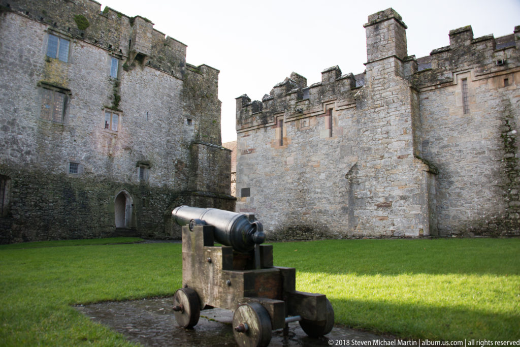 Dark cannon on a wheeled carriage in a grassy Cahir Castle courtyard beneath crenellated stone walls.