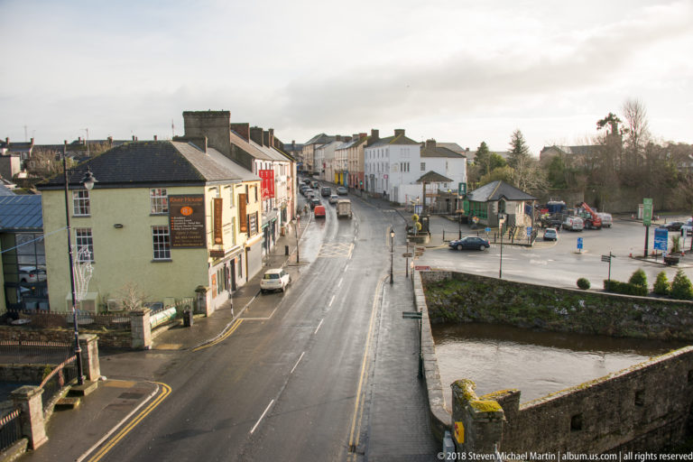 Elevated view from Cahir Castle over a wet town street, low buildings, parked cars, and the river edge.
