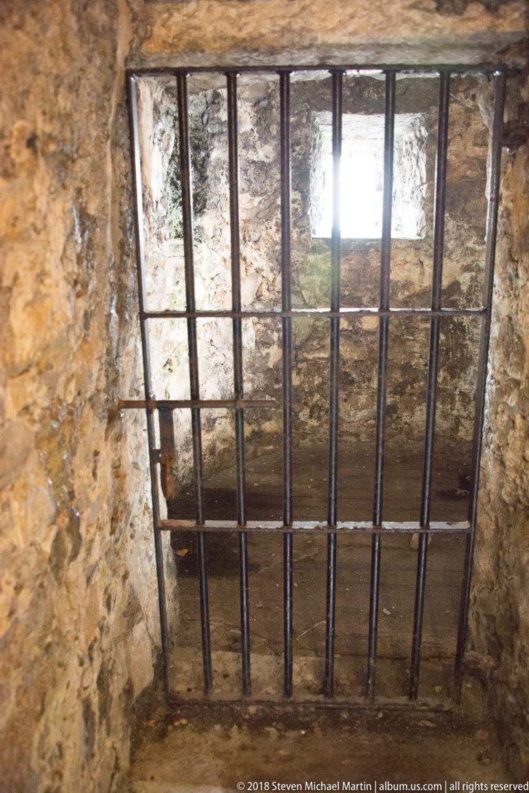 Iron grille with vertical and horizontal bars covering a narrow opening through rough castle masonry.