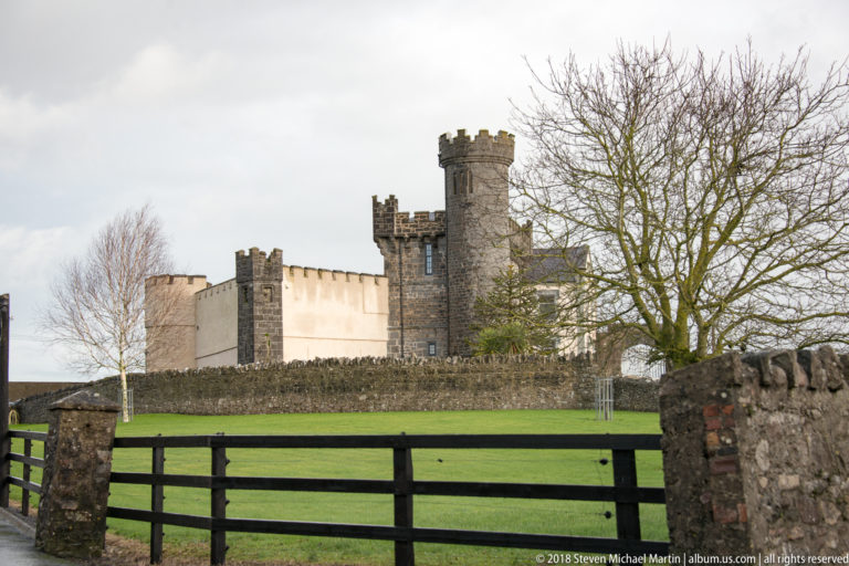 Cahir Castle towers and walls beyond a green lawn, boundary wall, metal gate, and leafless trees.