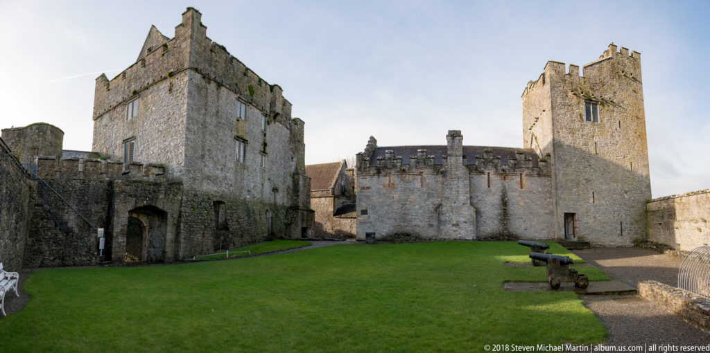 Grassy Cahir Castle courtyard enclosed by tall stone towers, with an arched doorway, benches, and cannon.