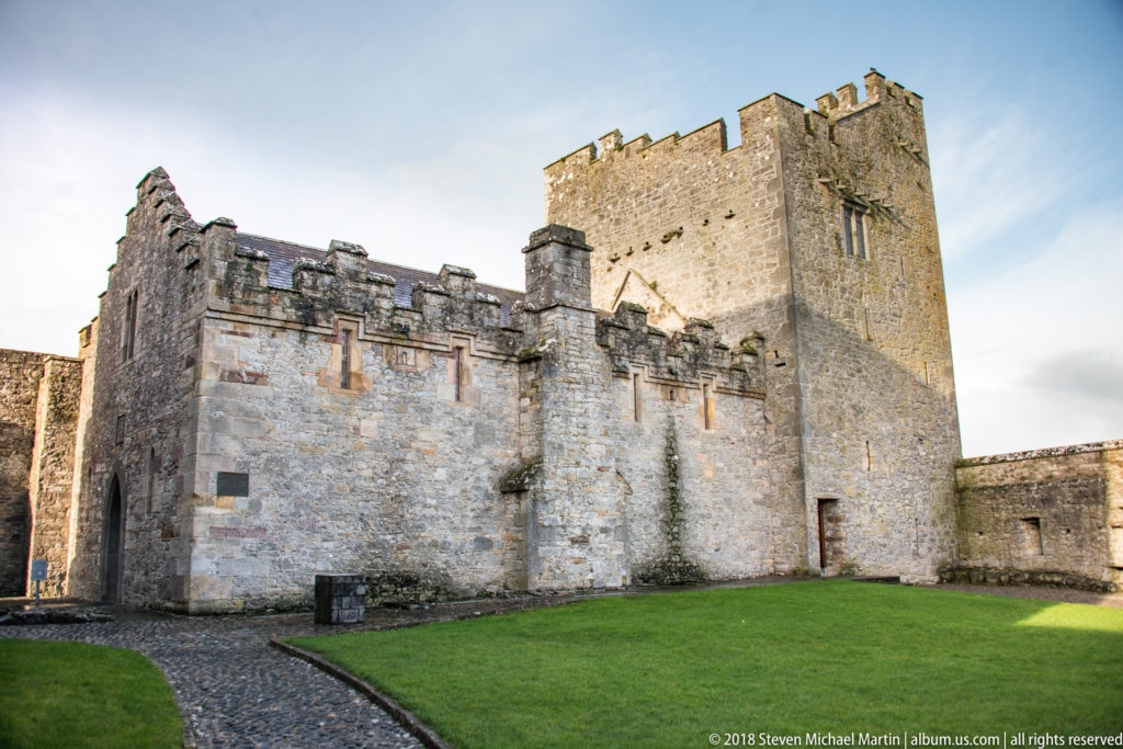 Square stone tower rising behind a lower crenellated wall beside a green lawn inside Cahir Castle.