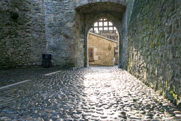 Rounded cobbles leading through a broad stone arch toward a sunlit courtyard and small building.