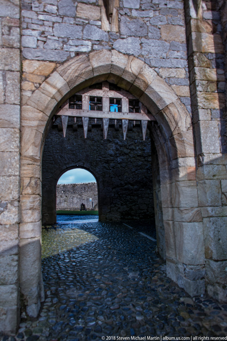 Raised wooden portcullis with pointed teeth above a stone archway leading through Cahir Castle.