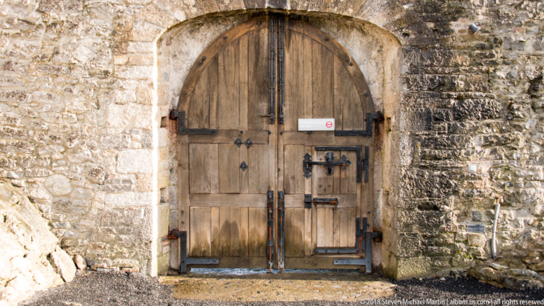 Heavy arched timber double doors with iron fittings and a smaller inset door in a weathered stone wall.