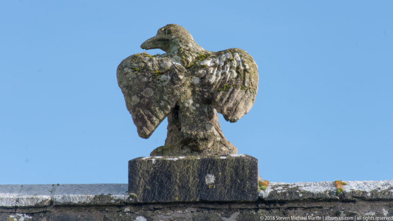 Weathered stone eagle with spread wings on a wall-top plinth against a clear blue sky.