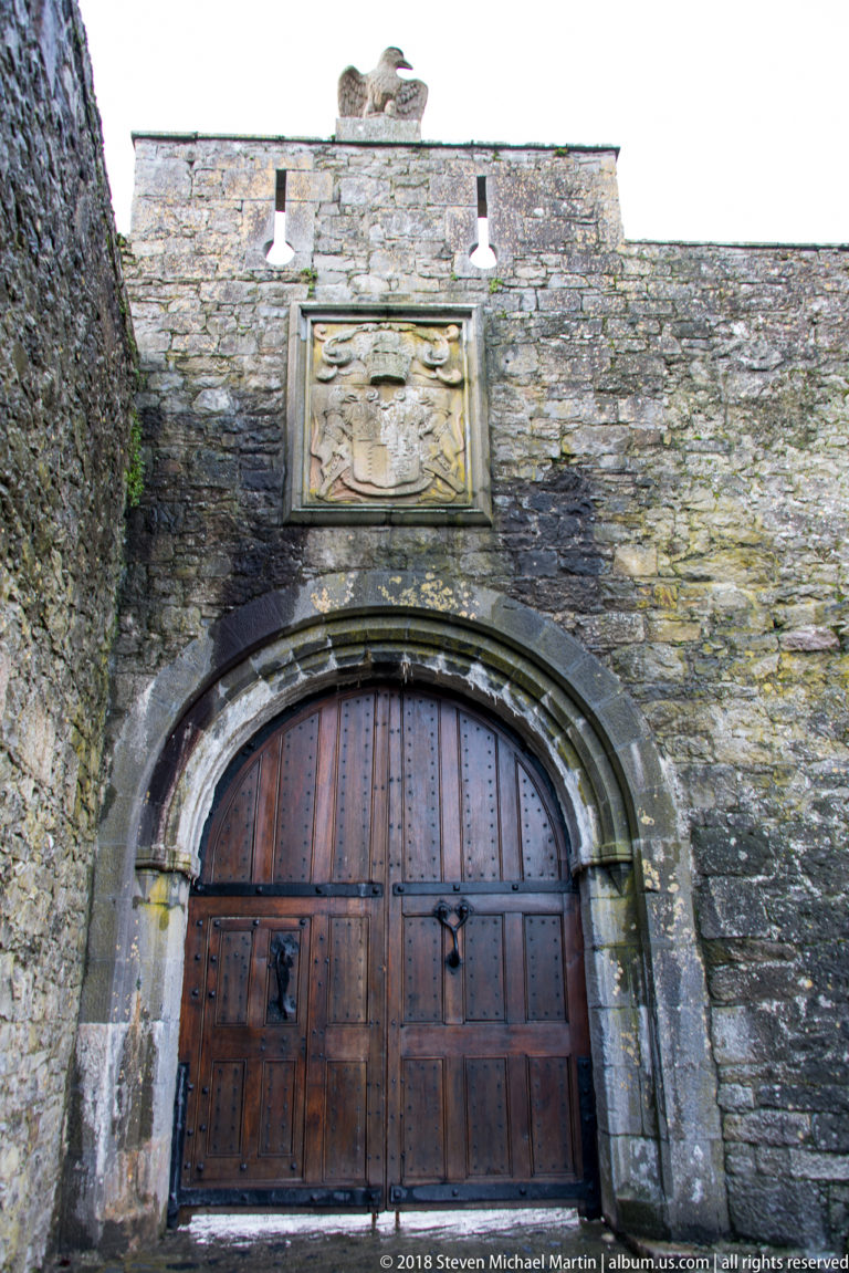 Large studded timber gateway in Cahir Castle beneath a carved heraldic panel and wall-top bird sculpture.