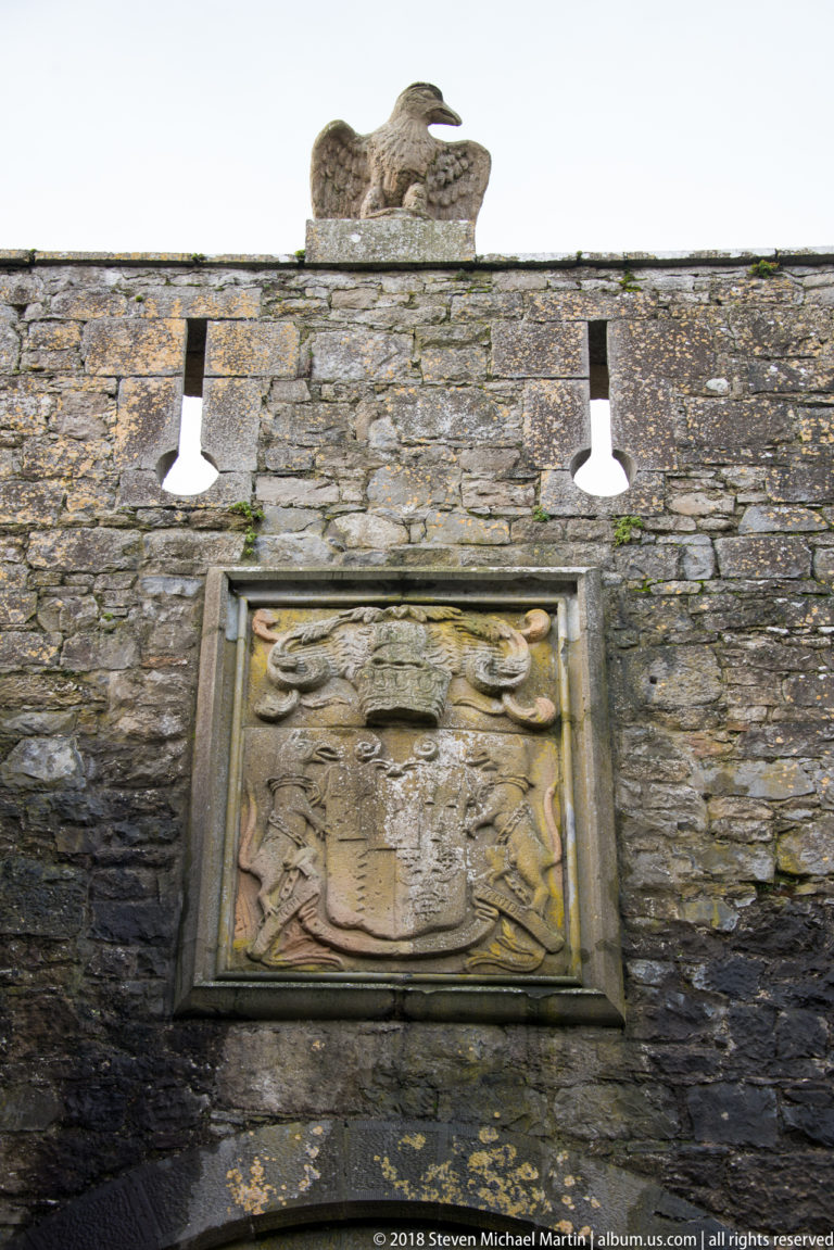 Eagle sculpture above a framed heraldic relief on a rough stone castle wall, with two modern lamps.