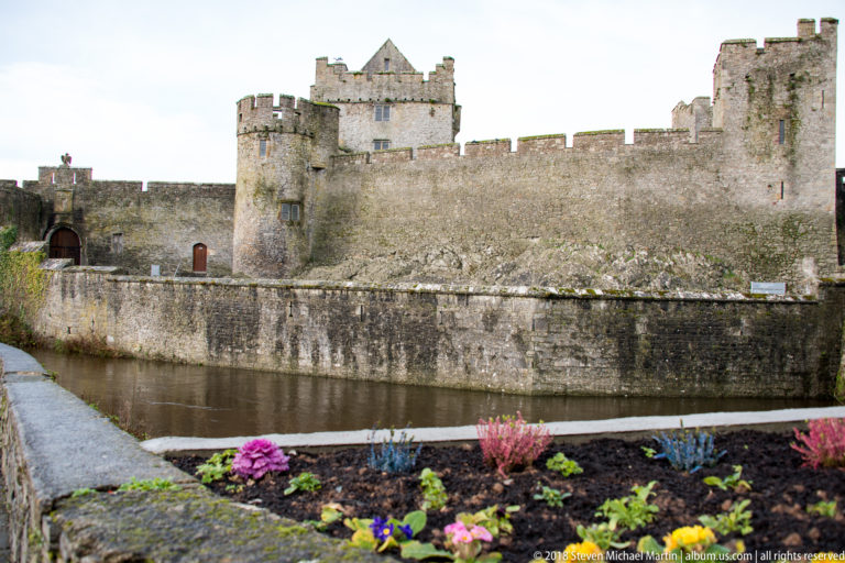 Cahir Castle’s crenellated stone walls and towers beside the river, with a flower bed in the foreground.
