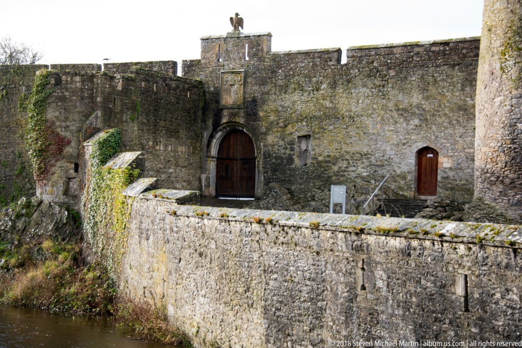 Arched wooden gateway in Cahir Castle’s stone wall, with heraldic panel above and river approach below.