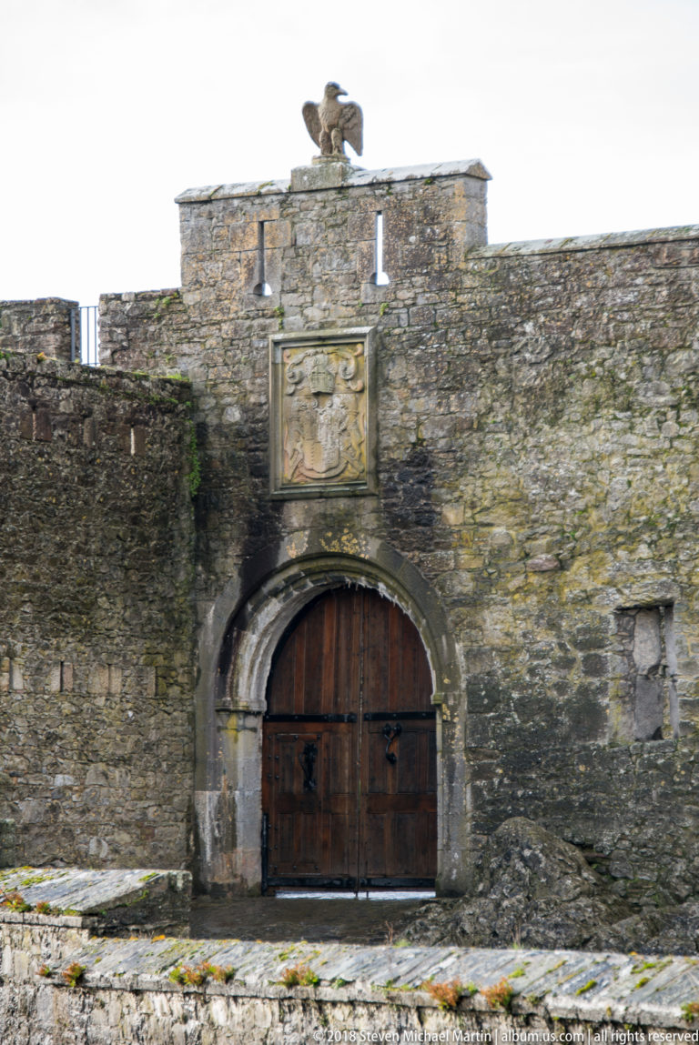 Closed wooden castle gate beneath a carved heraldic panel and bird sculpture on the parapet.