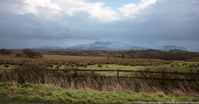 Distant snowy ridge beneath heavy clouds, seen across rough pasture, hedges, fencing, and scattered winter trees.