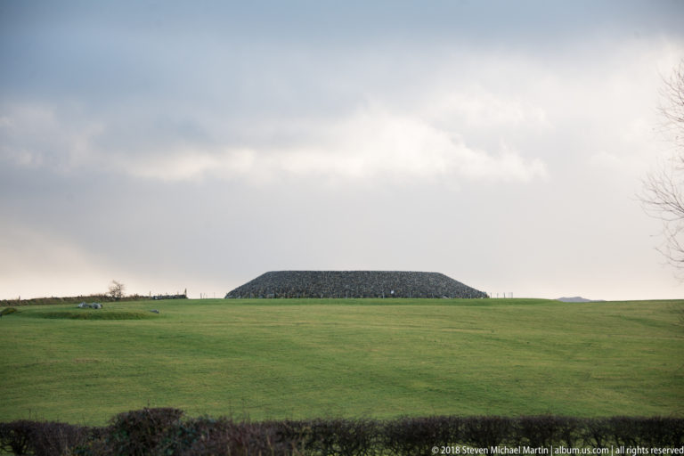 Large low stone cairn on the crest of a broad green field beneath a cloudy sky.