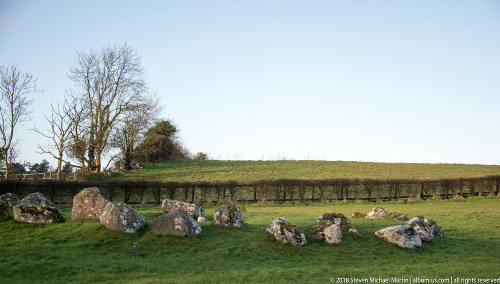 Low circular arrangement of weathered stones in green pasture, with hedgerows and leafless trees beyond.