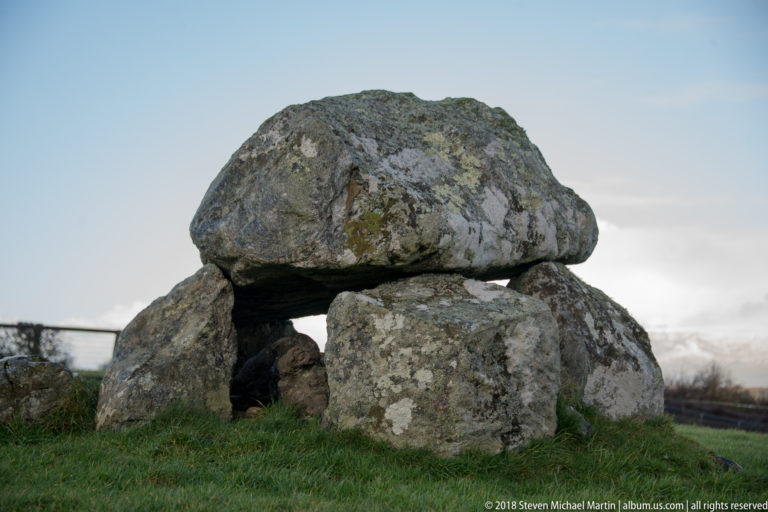 Broad capstone spanning upright stones above a deep dark chamber opening in a grassy field.