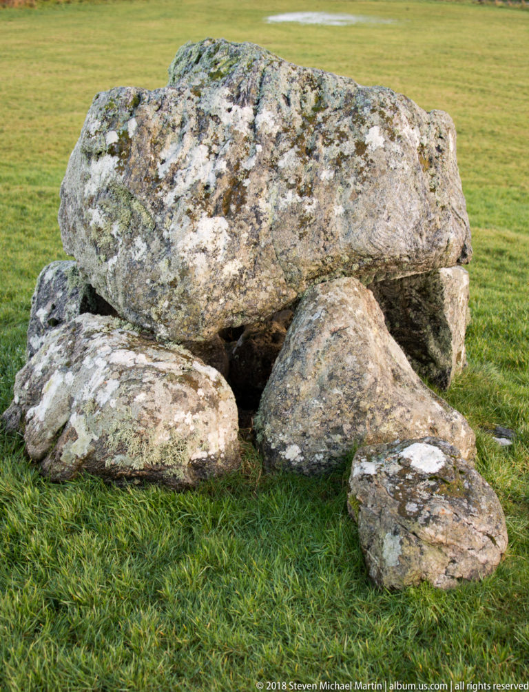 Close side view of a broad capstone resting over several large weathered stones with a dark recess beneath.