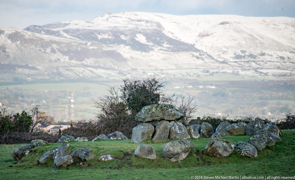 Megalithic tomb surrounded by a low ring of stones, with a snow-covered mountain in the background.