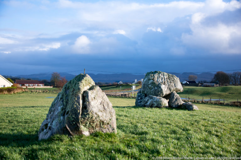 Large upright stone in the foreground with a low megalithic tomb farther across green pasture.