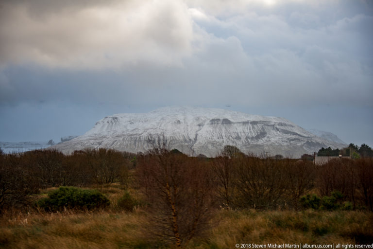 Closer view of a snow-covered flat-topped mountain with horizontal rock layers above leafless trees and scrub.