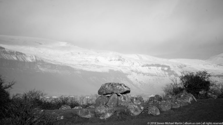Black-and-white view of a capstone tomb and scattered stones beneath a snow-covered mountain.