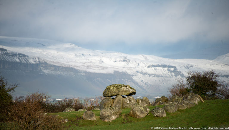 Megalithic tomb and surrounding stones in green pasture with a snow-covered mountain filling the background.