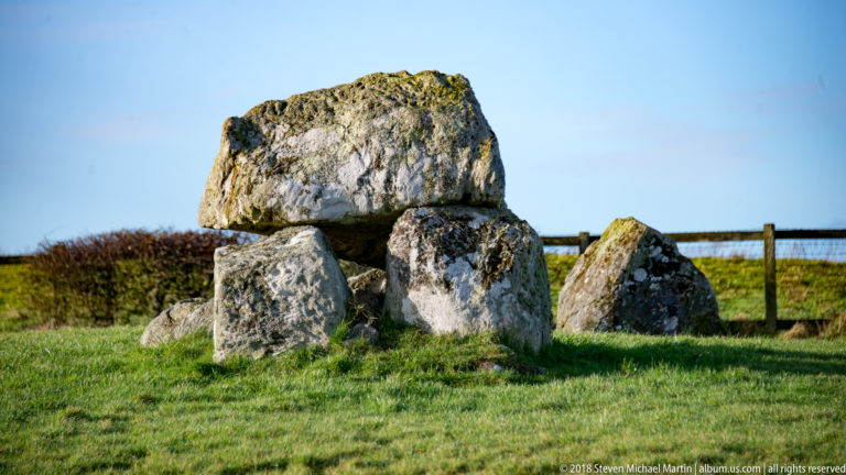 Compact megalithic tomb with a thick capstone on upright stones beside a wooden fence.