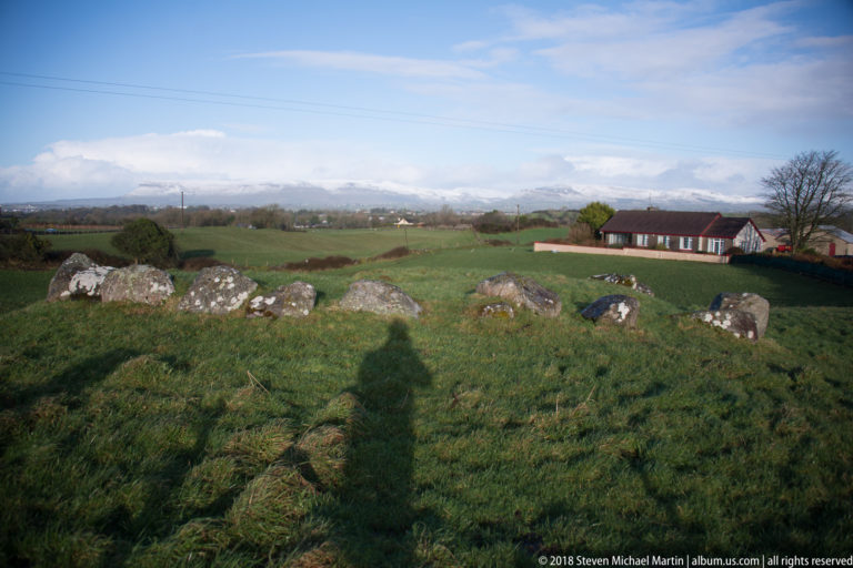 Low stone ring in pasture seen toward rural buildings, with the photographer’s shadow crossing the foreground.