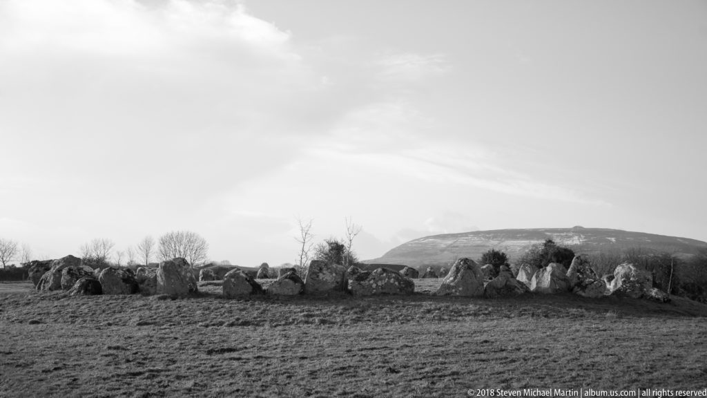 Black-and-white view of a low megalithic stone circle crossing open pasture beneath a rounded hill.
