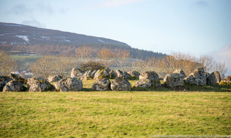 Low arc of large weathered stones in green pasture, backed by bare trees and a dark upland slope.