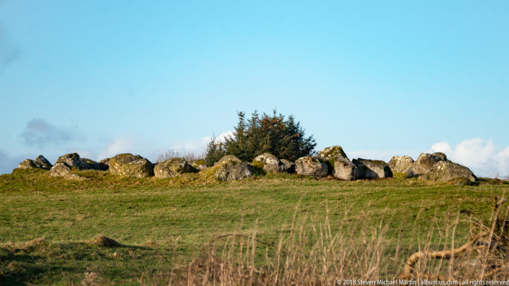 Irregular megalithic boulders forming a low line along a grassy ridge beneath blue sky.