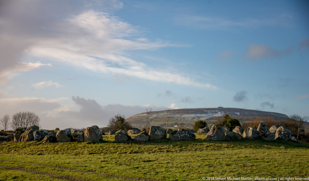 Low ring of megalithic stones crossing a grassy field, with a broad mountain in the distance.